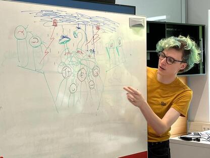 Myself, with green hair and a yellow shirt, leaning over whiteboard, explaining my team's illustration of people with glyphs on their face, tendrils intertwined between their heads, mushrooms growing from the tendrils, and a thunderstorm abvoe showering them all in water.